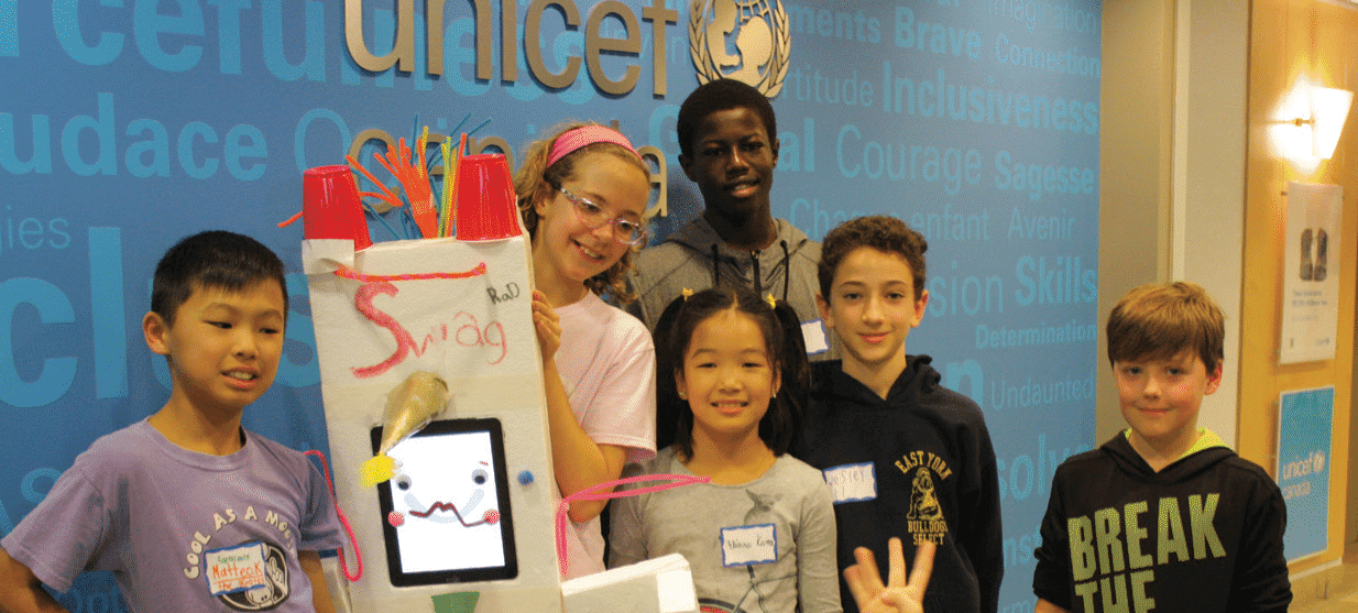 A diverse group of six children stand together, smiling, in front of a blue UNICEF Canada background. They are holding a creative project made from various materials, featuring an animated face on a screen and the word 'Swag' written on it. The setting suggests a workshop or event focused on youth engagement.