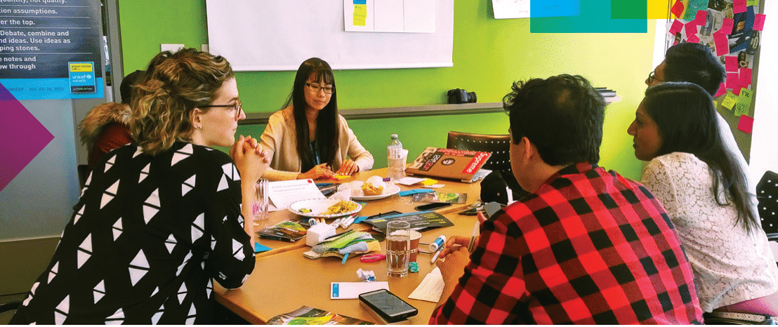 A group of young adults sits around a table in a discussion, with papers, booklets, and snacks spread out in front of them. The participants appear engaged in conversation, and one person listens attentively while others speak. The background features a green wall, sticky notes, and a poster with motivational text. The setting suggests a collaborative workshop focused on youth-adult engagement.