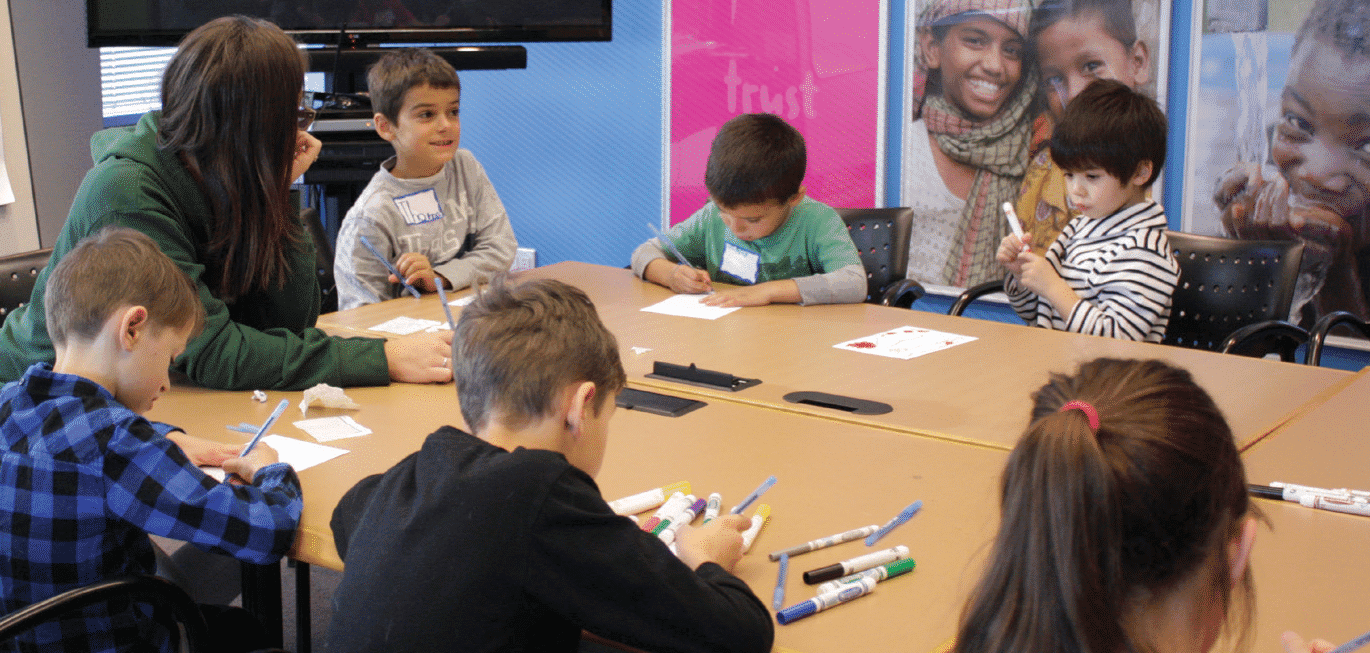 A group of young children sits around a table, engaged in drawing and coloring activities. An adult sits with them, guiding their work. The room has colorful posters in the background, with images of smiling children and words like 'Trust.' The setting appears to be a welcoming, educational space designed for youth engagement and development.
