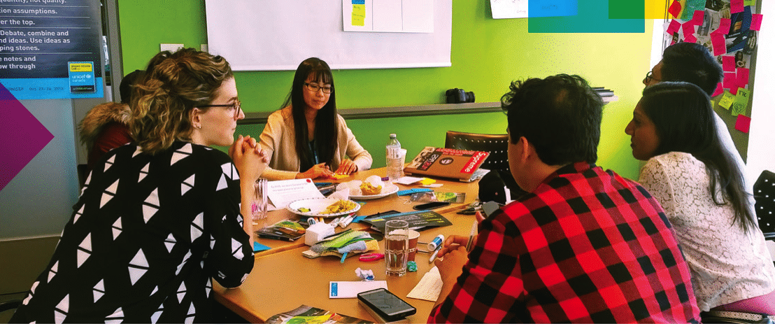 Un groupe de jeunes adultes est assis autour d'une table en pleine discussion, avec des papiers, des brochures et des collations étalés devant eux. Les participants semblent engagés dans la conversation : une personne écoute attentivement tandis que d'autres s'expriment. En arrière-plan, on voit un mur vert, des notes autocollantes et une affiche avec un texte motivant. Le cadre suggère un atelier collaboratif axé sur l'engagement des jeunes et des adultes.